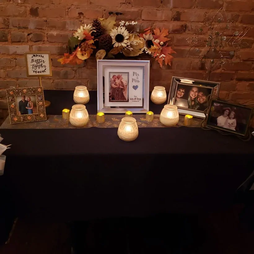 A candlelit memory table with framed photos and fall florals against brick at The Attic